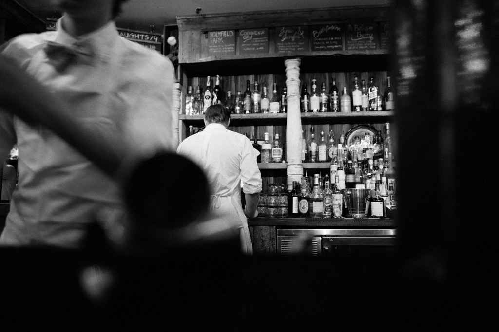 The bartender standing in front of a the bottles of alcohol.