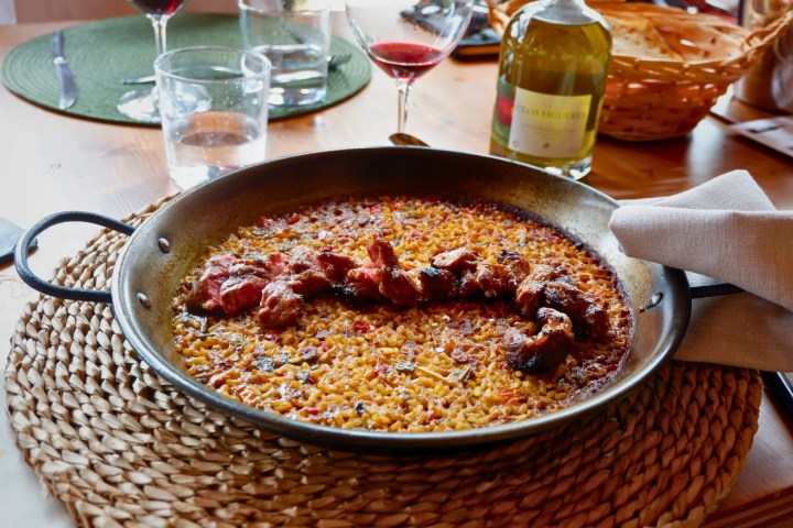 Paella with meat chunks in a pan on a woven mat, surrounded by glasses and a wine bottle.