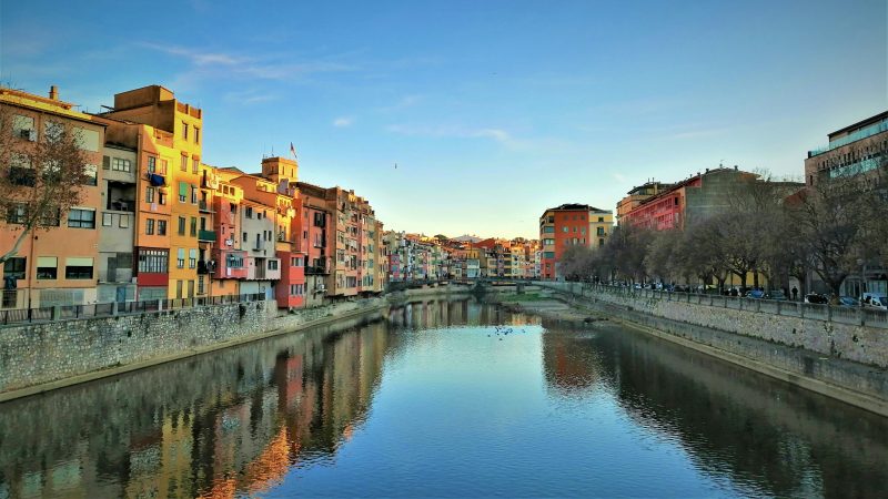 Colorful buildings and trees reflected in a calm river under a clear blue sky.