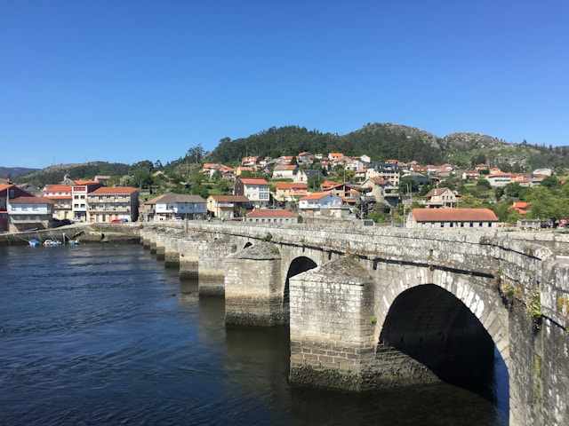 Stone bridge over river, houses on hill, clear blue sky.